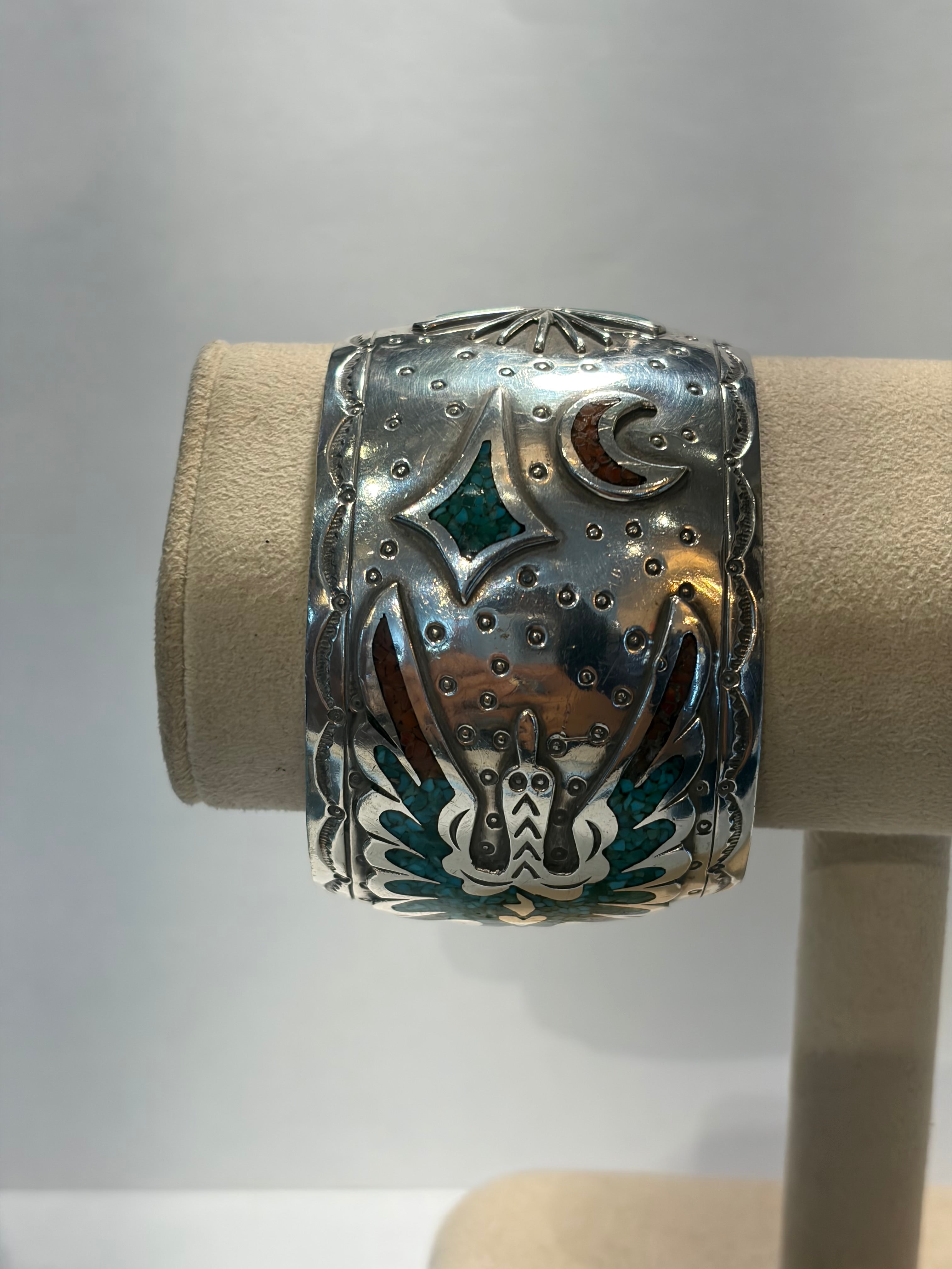 A wide silver cuff bracelet with intricate hand-set  turquoise and coralinlay details, displayed on a beige fabric jewelry stand.